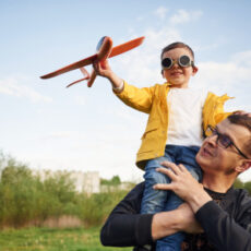 Boy is sitting on man's shoulders. Father with his little son playing with toy plane on the field.
