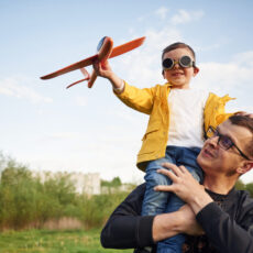 Boy is sitting on man's shoulders. Father with his little son playing with toy plane on the field
